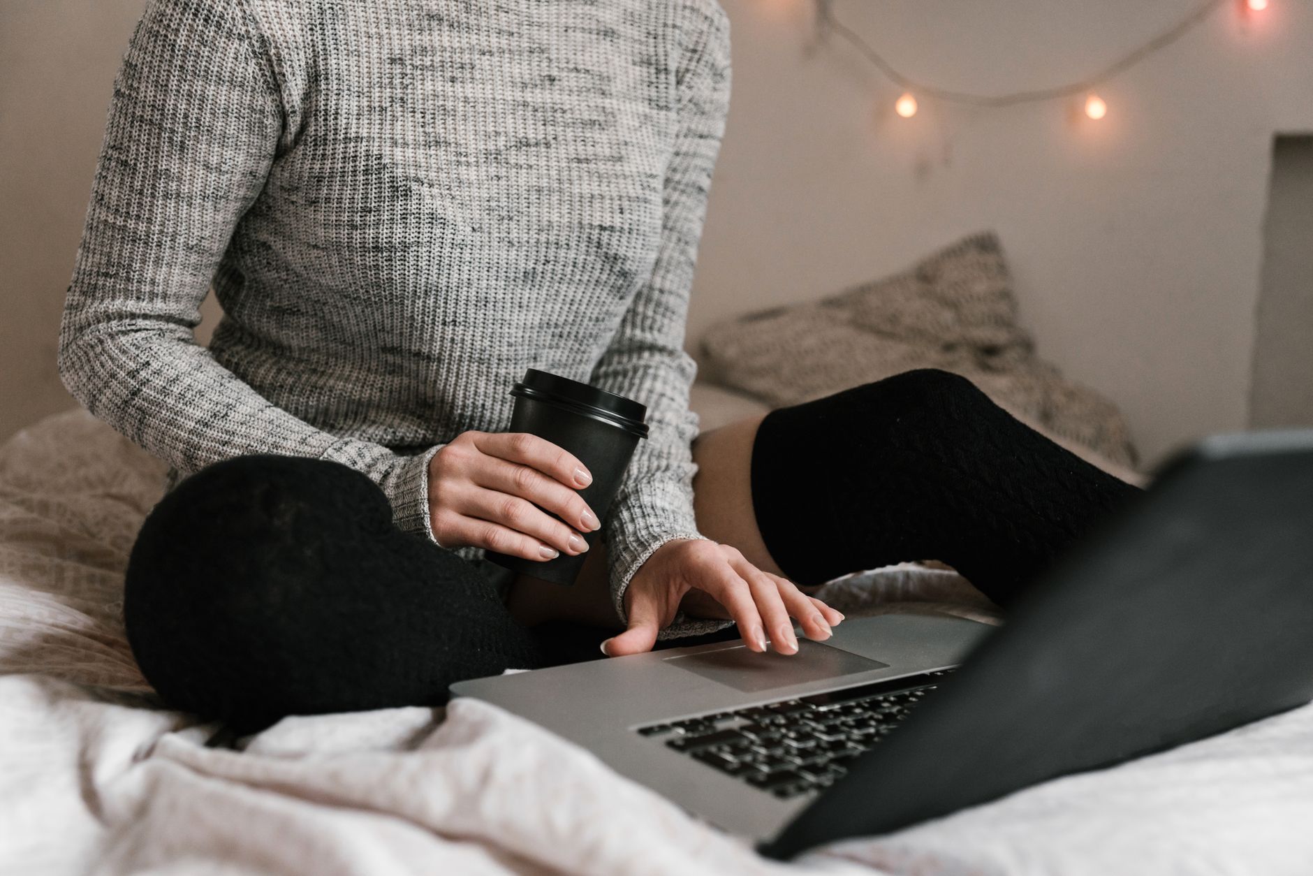 Une personne en pull avec un café et un ordinateur portable, installée sur un lit pour travailler tranquillement.